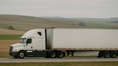 A conventional sleeper tractor coupled to a dry van trailer at an open interstate through rolling farmland